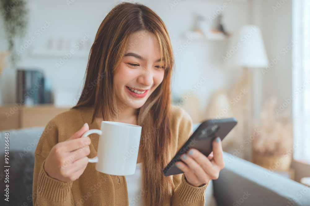 Relaxed young asian woman using mobile smartphone and drinking coffee from a mug while sitting on the sofa. Relaxed young asian woman using mobile smartphone and drinking coffee from a mug while sitting on the sofa.