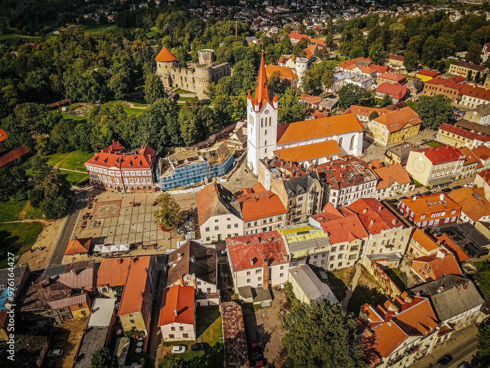 Obraz premium A vibrant aerial shot of a historic town, highlighting a central church with a red roof and tall steeple, alongside a medieval castle surrounded by lush green trees. The surrounding buildings feature