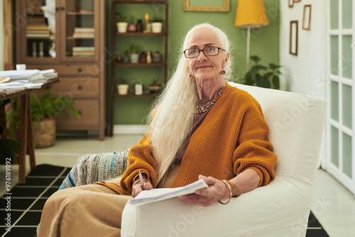 Portrait of elderly woman smiling while holding papers and sitting in stylish modern living room, showcasing a cozy and inviting space with elegant decor photo