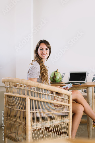 Portrait of cheerful woman manager of cozy Indonesian cafeteria sitting onbamboo chair and smiling at camera during organisation time enjoying own lifestyle and favourite job, positive digital nomad photo