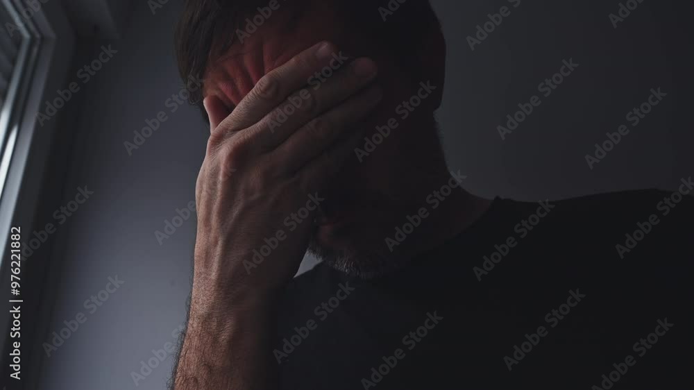 Sad and alone disappointed man crying in despair in his room with light and shadow coming from the window Sad and alone disappointed man crying in despair in his room with light and shadow coming from the window
