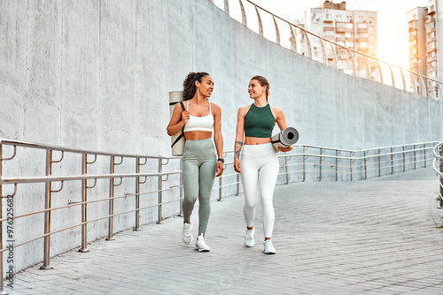 Two attractive modern sports women are walking holding mats for fitness training, yoga and joyfully chatting. Sincere friendship and sports lifestyle.Copy space.