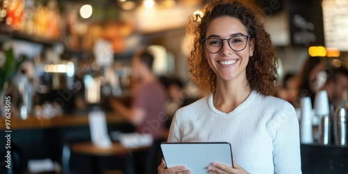 A woman wearing glasses and a white sweater stands in a bustling cafe holding a tablet and smiling, embodying a friendly and approachable ambiance.