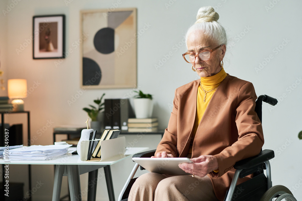 Elderly woman wearing glasses and business attire holding tablet while sitting in modern office. Background features contemporary art and neatly arranged office supplies Elderly woman wearing glasses and business attire holding tablet while sitting in modern office. Background features contemporary art and neatly arranged office supplies