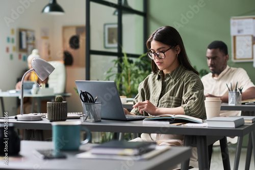 Woman in green shirt typing on laptop in modern shared workspace surrounded by colleagues, books, and plants creating collaborative environment multiple working professionals