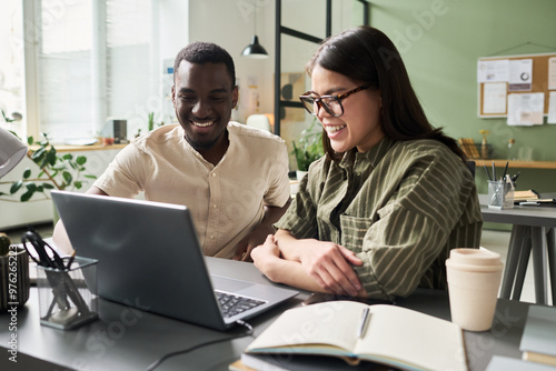 Colleagues working together, smiling, and looking at computer screen in modern office setting with desk and coffee cup. Lively interaction between coworkers, enhancing productivity