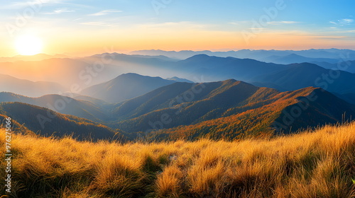 Landscape of majestic mountain ranges at sunrise, with golden sunlight casting soft rays over distant hills and valleys. The foreground consists of a grassy ridge covered in golden autumn hues.