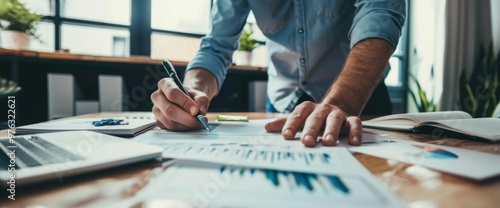 A businessman in a casual shirt analyzes financial reports on a desk, with a laptop and documents, in a modern office setting.