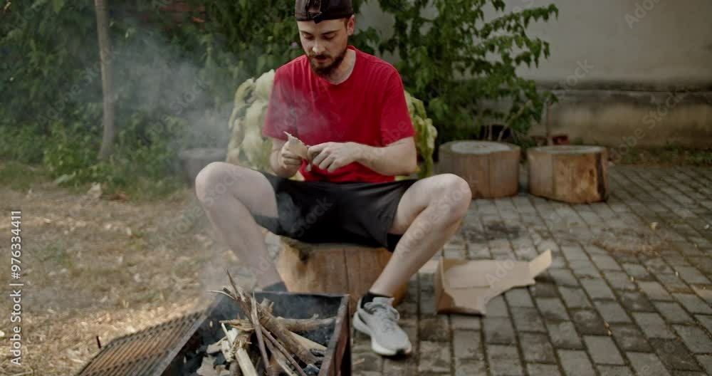 A man in a red shirt and black shorts prepares the wood and charcoal in a barbecue in a yard. His friend watches on.