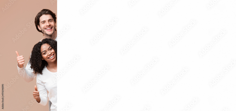 Happy multiracial couple peeking behind white advertisement board and showing thumbs up, recommending something Happy multiracial couple peeking behind white advertisement board and showing thumbs up, recommending something