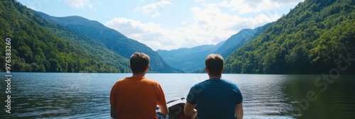 Two individuals, seen from the back, enjoy a peaceful moment by the lakeside, surrounded by lush green mountains under a bright blue sky, reflecting calmness and tranquility. photo