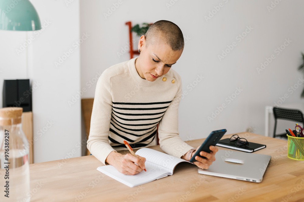 Mid adult woman taking notes while checking some information on mobile phone sitting at workplace in the office. Technology concept. Mid adult woman taking notes while checking some information on mobile phone sitting at workplace in the office. Technology concept.