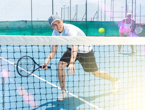 Portrait of emotional young guy playing tennis on open court in summer, swinging racket to return ball over net. Sportsman ready to hit volley photo