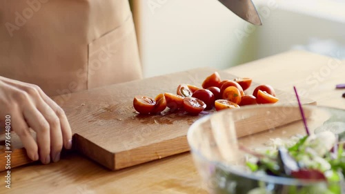 Woman preparing vegetable salad, cutting and adding cherry tomatoes into bowl, close up