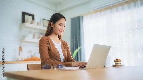 Beautiful young Asian woman working with laptop computer in home kitchen. Work at home