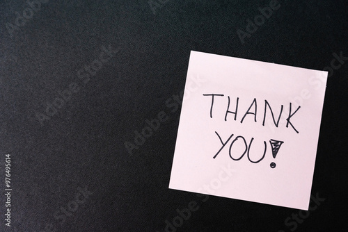 Hand writing THANK YOU on pink sticky note isolated on dark background. Copy space.