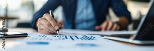 In a professional office setting, a person is focused on writing on documents with a pen, surrounded by blue graphs, charts, and a laptop, indicating work and analysis. photo