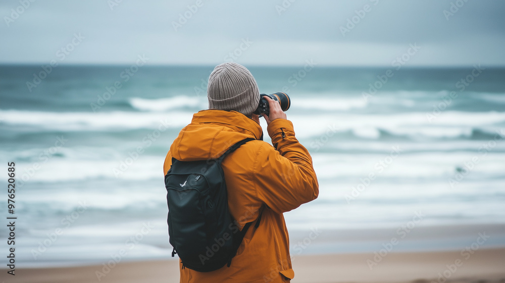 A male photographer with a beanie and a backpack takes photos by the beach on a cloudy day. A male photographer with a beanie and a backpack takes photos by the beach on a cloudy day.