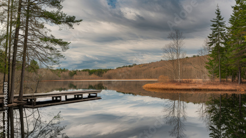 A photo of a serene lake surrounded by tall trees, with a wooden dock extending into the water. The reflection of the trees and dock is mirrored on the lake's calm surface. The sky is overcast