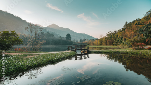 A photo of a serene landscape with a clear sky. There's a small wooden bridge over a calm lake. The lake reflects the sky and the bridge. The surrounding area is filled with lush greenery, with trees 