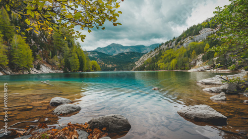 A photo of a serene lake with crystal clear water, surrounded by lush green forests and rocky terrain. The background contains a few trees and a few mountains with a cloudy sky.