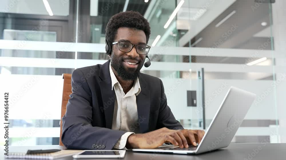Portrait of happy African American businessman in headset sitting at workplace in business office. Smiling manager or corporate operator in formal suit working at desk with laptop looking at camera Portrait of happy African American businessman in headset sitting at workplace in business office. Smiling manager or corporate operator in formal suit working at desk with laptop looking at camera