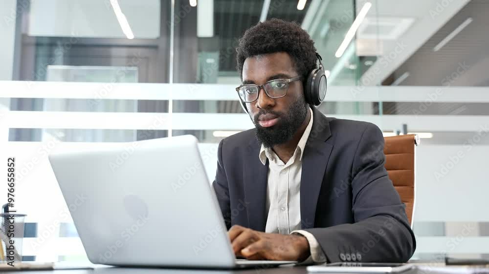 Confident african american IT specialist developer in wireless headphones works on laptop computer at workplace in business office. Serious focused coder in glasses writing code, working on project Confident african american IT specialist developer in wireless headphones works on laptop computer at workplace in business office. Serious focused coder in glasses writing code, working on project
