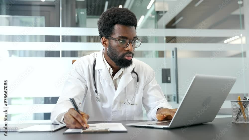 Focused African American doctor working at desk with laptop and documents in an office while wearing a white coat and stethoscope. Medical worker physician takes notes in a modern hospital clinic Focused African American doctor working at desk with laptop and documents in an office while wearing a white coat and stethoscope. Medical worker physician takes notes in a modern hospital clinic