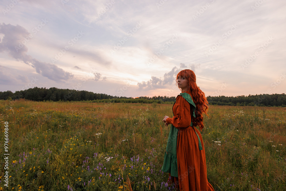 Fototapeta premium Beautiful woman with red hair in long red and green dress standing on flower field. Cloudy sky at sunset. Concept of rural romance, rural scene or love story.
