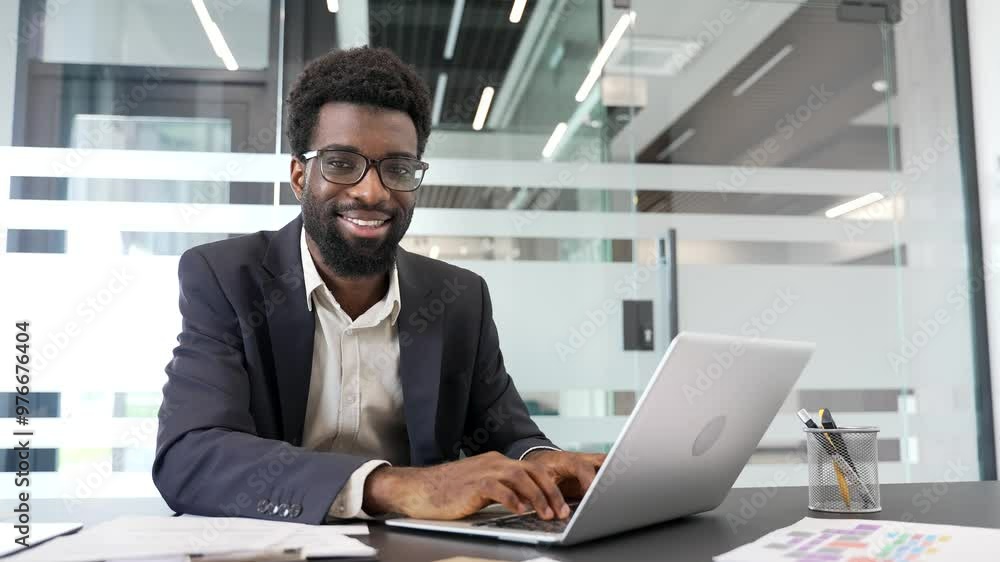 Portrait of happy African American businessman sitting at workplace in business office. Head shot of smiling handsome manager in formal suit working at desk with laptop computer and looking at camera Portrait of happy African American businessman sitting at workplace in business office. Head shot of smiling handsome manager in formal suit working at desk with laptop computer and looking at camera