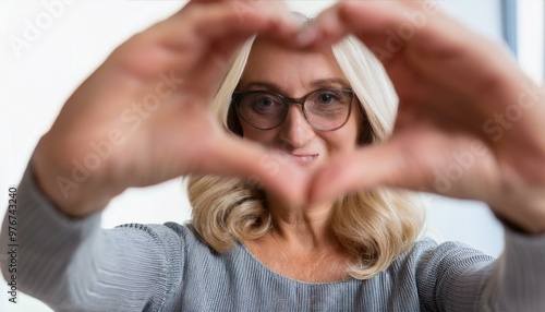 portrait of a person with a headache,Mature woman wears eyeglasses looking at camera through joined fingers showing heart symbol,