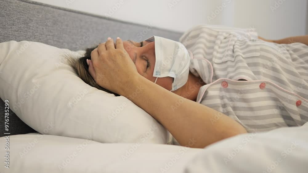 Sick woman wearing mask lying in bed fighting illness. Visible discomfort from virus, cold, fever, cold and fatigue from illness. Lying in bed due to severe illness showing the impact of illness. Sick woman wearing mask lying in bed fighting illness. Visible discomfort from virus, cold, fever, cold and fatigue from illness. Lying in bed due to severe illness showing the impact of illness.
