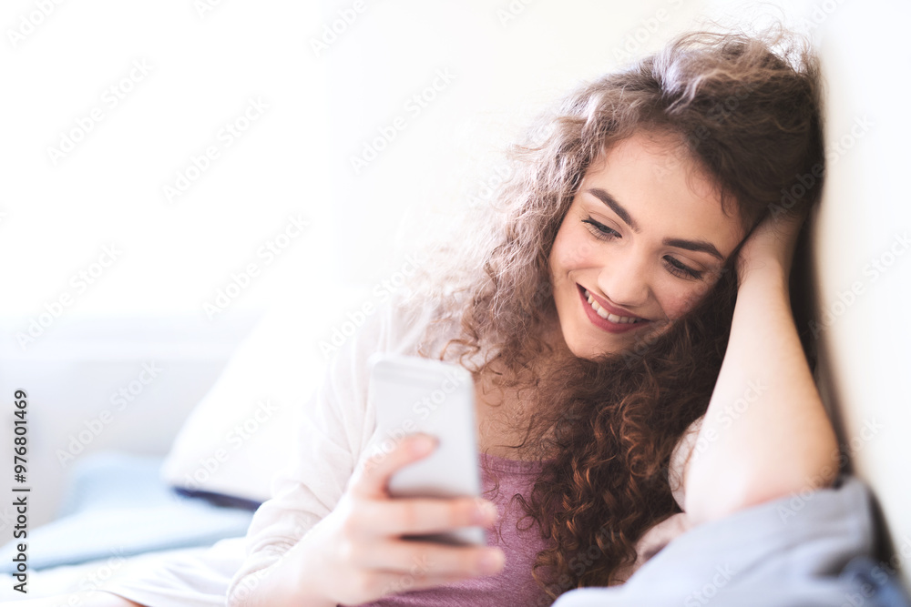 Beautiful young woman sitting in bed, scrolling on smartphone, smiling. Beautiful young woman sitting in bed, scrolling on smartphone, smiling.