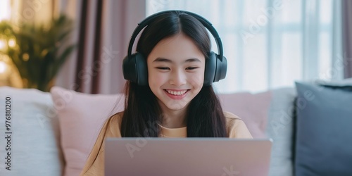 A young girl is sitting on a couch with a laptop in front of her. She is wearing headphones and smiling photo