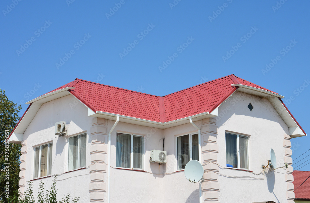 House with red lightweight metal roofing sheets, white plastic rain gutters, tv antenna, air duct and white painted walls against blue sky House with red lightweight metal roofing sheets, white plastic rain gutters, tv antenna, air duct and white painted walls against blue sky