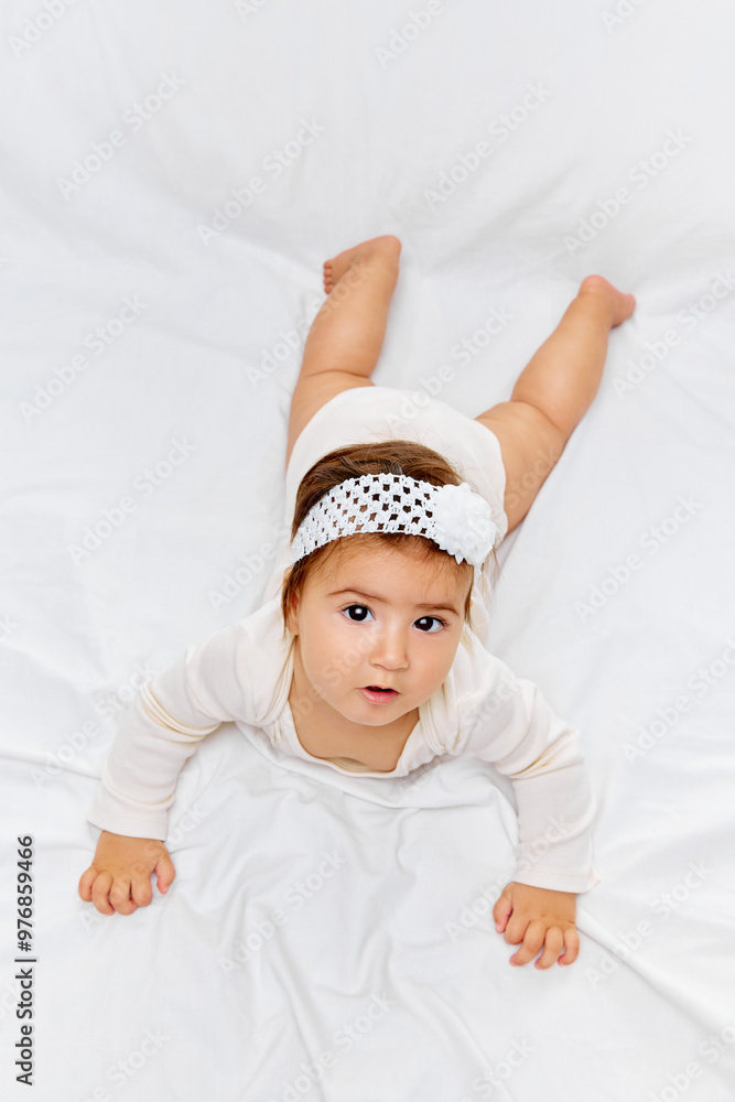 Young child in white onesie lies flat on her stomach, gazing intently at camera against white studio background. Concept of beauty, parenthood, childhood, life and birth. Young child in white onesie lies flat on her stomach, gazing intently at camera against white studio background. Concept of beauty, parenthood, childhood, life and birth.