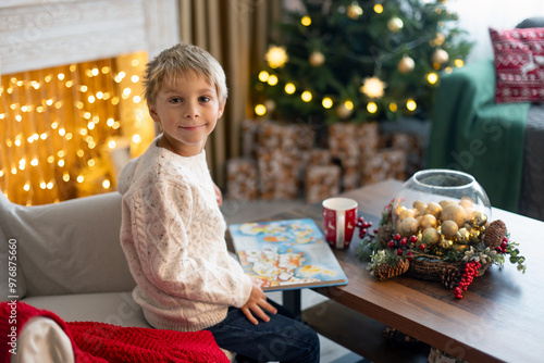 Cute blond child, boy, celebrating Christmas at home, decorated room for the holidays, child reading book and playing photo