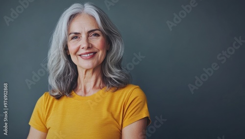 Portrait of a beautiful mature woman with gray hair, wearing a yellow T-shirt and looking at the camera against a gray background. This concept image could be used for skin care and beauty treatment 