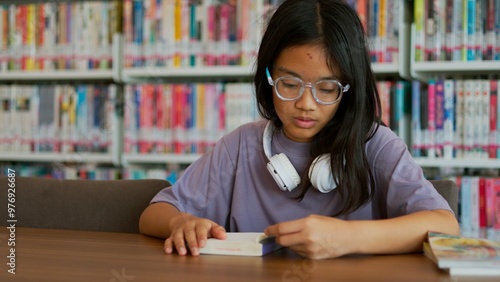 An Asian woman sits in a library, focused intently on a book, her expression serious.