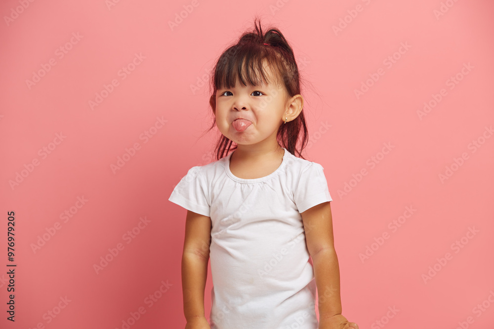 Little Asian baby girl shows tongue wears t shirt isolated over pink background. Three years old offended and capricious child. Little Asian baby girl shows tongue wears t shirt isolated over pink background. Three years old offended and capricious child.