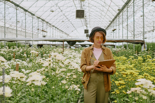 Obraz na plátně Woman standing among blooming flowers in large greenhouse, wearing hat and glass