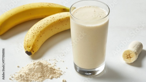 A glass of vanilla protein shake next to a banana and a scoop of protein powder on a white surface.