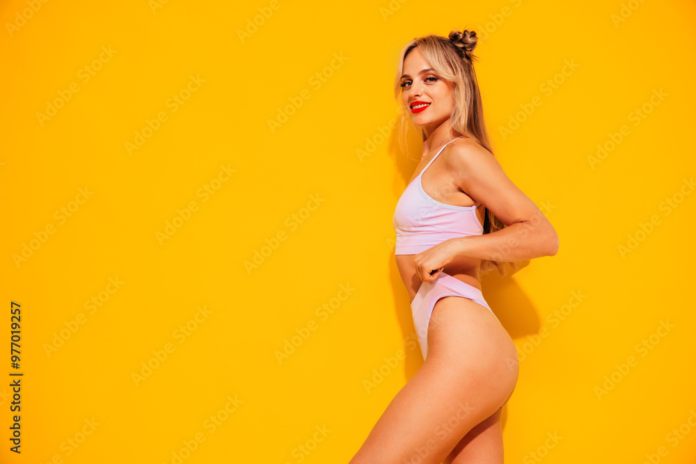 Young smiling blond model in summer swimwear colourful bathing suit. Sexy carefree woman having fun and going crazy. Female posing near yellow wall in studio. Cheerful and happy Young smiling blond model in summer swimwear colourful bathing suit. Sexy carefree woman having fun and going crazy. Female posing near yellow wall in studio. Cheerful and happy
