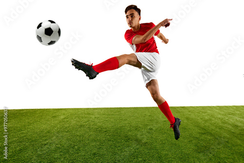 Young athletic man, football player, dressed in red and white uniform, performs high kick, focusing on controlling ball with precision on green field. Concept of championship, sport events, final game