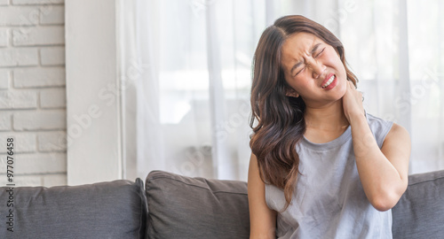Asian woman sitting on sofa with a painful expression holding her neck, suffering from neck pain indoor home setting, casual clothing, woman health issue, discomfort, stress, fatigue at living room Free Download Asian woman sitting on sofa with a painful expression holding her neck, suffering from neck pain indoor home setting, casual clothing, woman health issue, discomfort, stress, fatigue at living room - GFXLoad