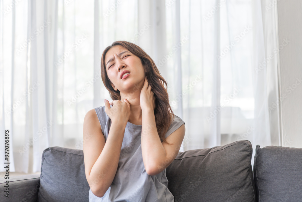 Asian woman sitting on sofa with a painful expression holding her neck, suffering from neck pain indoor home setting, casual clothing, woman health issue, discomfort, stress, fatigue at living room