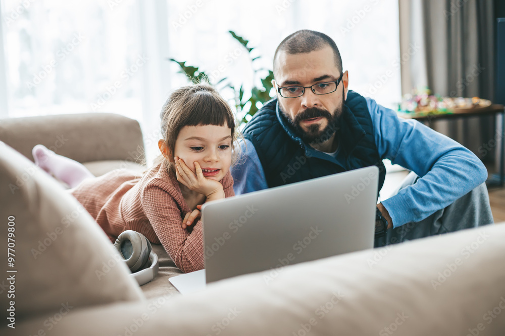 A father and his little daughter share a collaborative moment on the couch, focusing together on a laptop screen, potentially for study or leisure A father and his little daughter share a collaborative moment on the couch, focusing together on a laptop screen, potentially for study or leisure