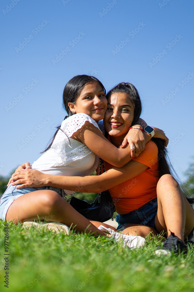 Portrait of two Latina sisters hugging each other sitting in a park, one teenager and one young adult Portrait of two Latina sisters hugging each other sitting in a park, one teenager and one young adult