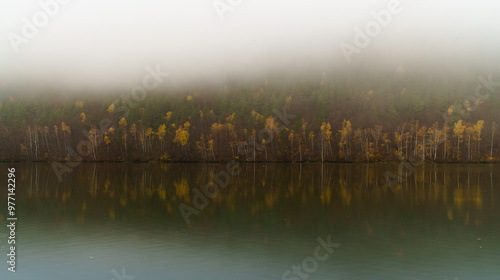 Wide Vag river calm autumnal waters with reflected yellow and red deciduous misty forest near Sutovo settlement, Slovak Republic. Moody weather and traveling concept. photo