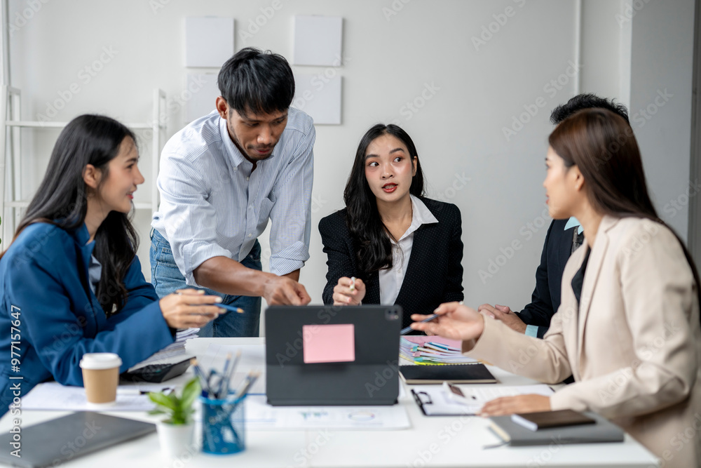 A group of people are sitting around a table, with a man pointing at a laptop A group of people are sitting around a table, with a man pointing at a laptop